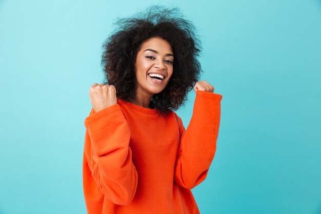 Il ritratto variopinto della donna felice in camicia rossa che guarda con il sorriso e che stringe i pugni gradisce il vincitore, isolato sopra la parete blu