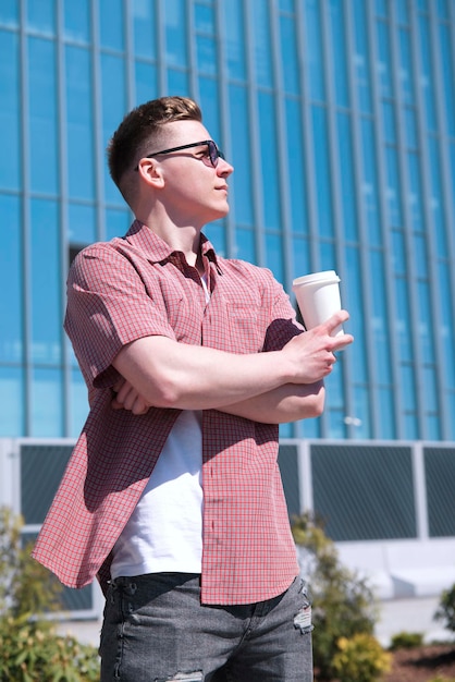 Il ritratto verticale del giovane bello alla moda sta bevendo il caffè o il tè dalla tazza di carta all'aperto a