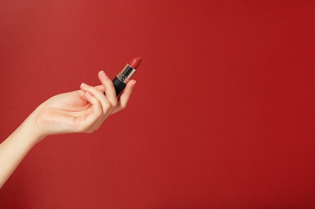 Il rossetto rosso in un tubo nero tiene una mano femminile su uno sfondo rosso. Prodotto cosmetico per il trucco creativo di bellezza brillante. Strumento per truccatori.