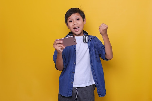 Il simpatico bambino asiatico in stile denim casual sta vincendo il gesto e sorride alla telecamera mentre tiene in mano lo smartph