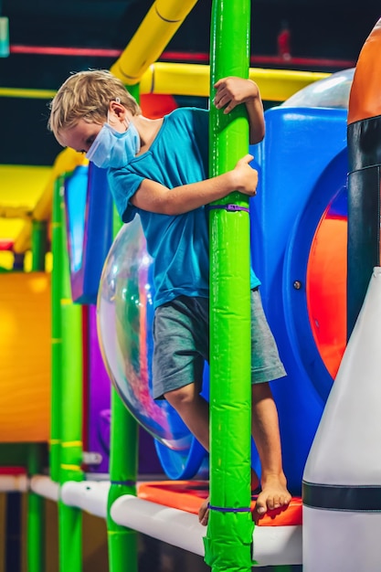 Il simpatico ragazzino biondo vuole divertirsi al colorato parco giochi al coperto del parco divertimenti sembra triste perché indossa una maschera protettiva blu nuovo stile di vita normale a causa dei cambiamenti in attesa di COVID19 Coronavirus