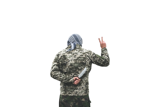Il soldato è in piedi in uniforme mimetica isolato su sfondo bianco. Bandana a scacchi in kefiah shemagh. Uomo con coltello dietro la schiena nel luogo deserto abbandonato. Tradimento dei nemici.