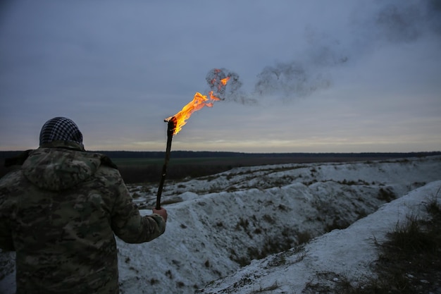 Il soldato tiene la torcia con il fuoco Uomo in uniforme mimetica e bandana a scacchi Persona nella gola delle montagne Colline bianche ed erba secca nel deserto