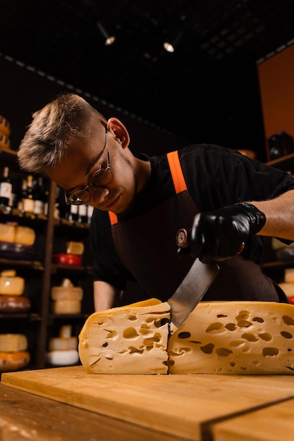 Il sommelier del formaggio nel negozio di alimentari sta tagliando maasdam invecchiato naturale e controlla la qualità Bell'uomo barbuto che lavora nel negozio di alimentari