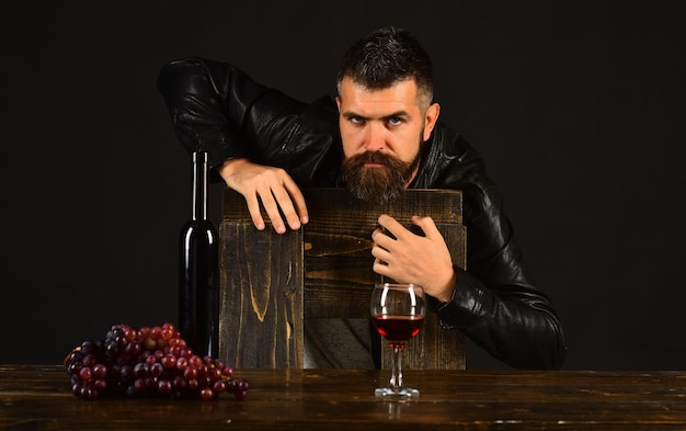 Il sommelier si appoggia sulla sedia di legno Uomo con la barba vicino a un bicchiere di vino su sfondo marrone scuro Degustatore con faccia seria da bottiglia di vino e uva scura Degustazione di vini e concetto di degustazione