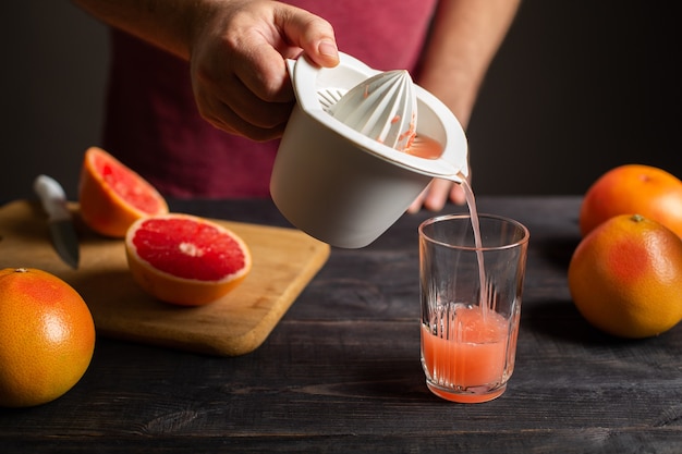 Il succo di pompelmo appena spremuto viene versato da uno spremiagrumi manuale in un bicchiere. Spremiagrumi in plastica bianca in mano maschio. Su un tavolo di legno nero ci sono pompelmi interi e affettati.