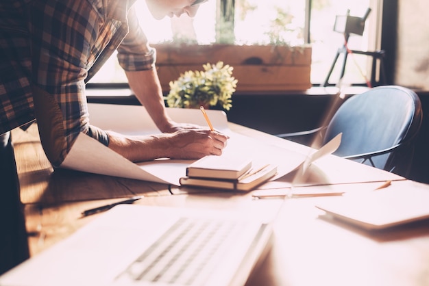 Il suo lavoro è la sua vita. Primo piano di un giovane fiducioso in abbigliamento casual che disegna sul progetto mentre si trova vicino alla scrivania di legno in un ufficio creativo