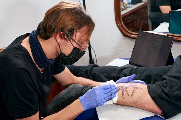 Il tatuatore in guanti blu che pulisce il tatuaggio appena completato sulla gamba di un uomo