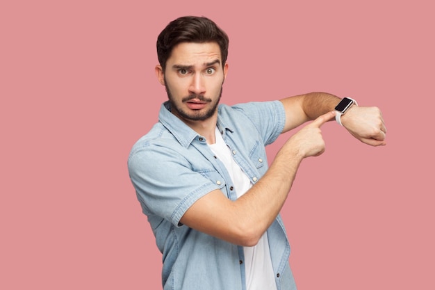 Il tempo è scaduto. Ritratto di giovane uomo barbuto bello serio in camicia blu in stile casual in piedi e guardando la fotocamera, puntando sul suo orologio intelligente. girato in studio al coperto, isolato su sfondo rosa.