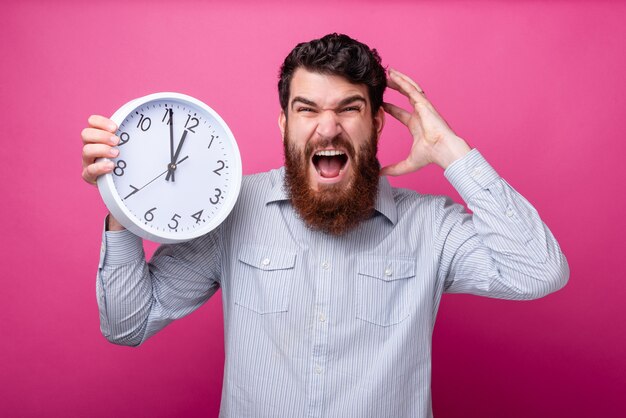 Il tempo scorre! Sono in ritardo. Il giovane uomo barbuto è stressato, tiene un orologio bianco e una mano sulla testa, urlando pazzo.