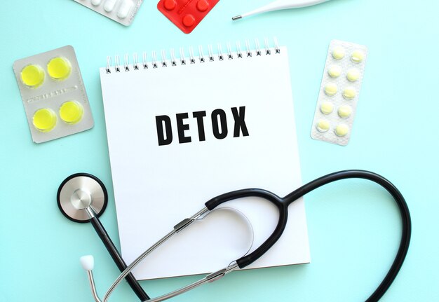 Il testo DETOX è scritto su un blocco note bianco che si trova accanto allo stetoscopio e alle pillole su uno sfondo blu.