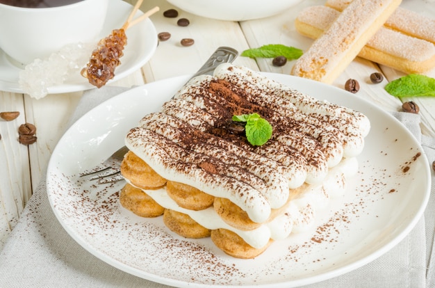 Il tiramisù italiano tradizionale del dessert è servito su un piatto con una tazza di caffè su una superficie di legno bianca