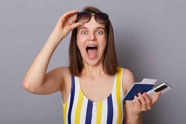 Il tiro al coperto di una donna estremamente felice e gioiosa che indossa il costume da bagno con in mano il passaporto ha sollevato i suoi occhiali da sole guardando la fotocamera urlando di felicità viaggiando isolato su sfondo grigio