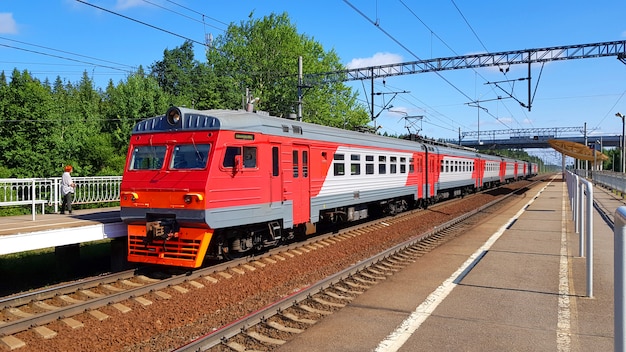 Il treno suburbano arriva alla stazione di estate il giorno soleggiato. Piattaforma ferroviaria con treno lungo il percorso.