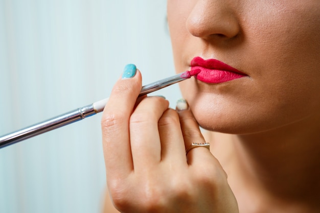 Il truccatore applica il rossetto rosso. Bel volto femminile. Mano di un maestro di trucco che dipinge le labbra di un modello di bellezza della ragazza. Trucco in corso