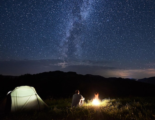 Il turista si sta rilassando vicino al falò godendosi le sagome delle montagne sotto il cielo stellato con la luminosa Via Lattea