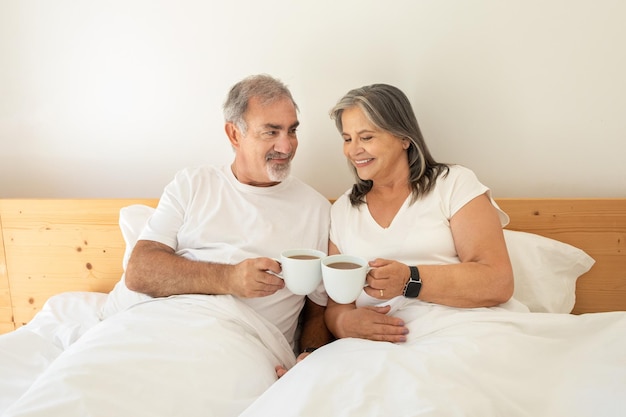 Il vecchio marito e la moglie caucasici sorridenti si trovano a letto e acclamano con tazze di caffè e si godono il buongiorno