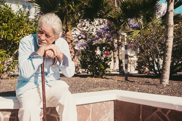 Il vecchio uomo anziano con le mani sopra il bastone seduto in un parco pubblico con espressione triste. Pensionato anziano con mal di schiena usando il bastone da passeggio. Albero e fiori sullo sfondo