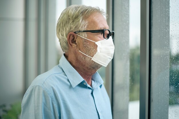 Il vecchio uomo caucasico indossa una maschera medica, guardando fuori dalla sua casa