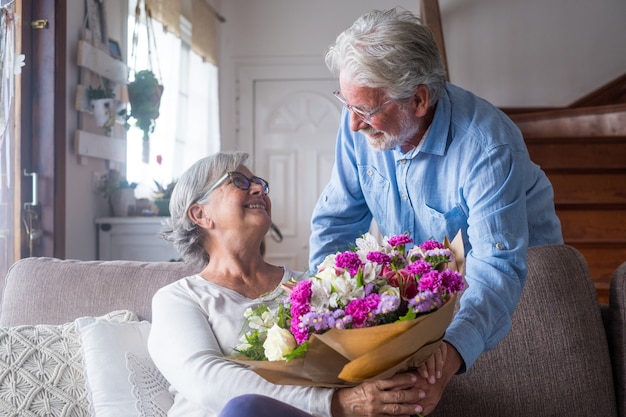 Il vecchio uomo che dà fiori a sua moglie seduta sul divano di casa per il giorno di San ValentinesÃ¢Â€Â™. Pensionati che si godono la sorpresa insieme. In amore le persone si divertono.