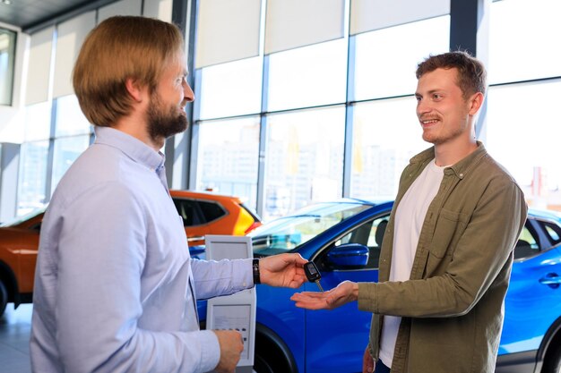 Il venditore della concessionaria di auto consegna la chiave dell'auto all'acquirente maschio Sullo sfondo della concessionaria