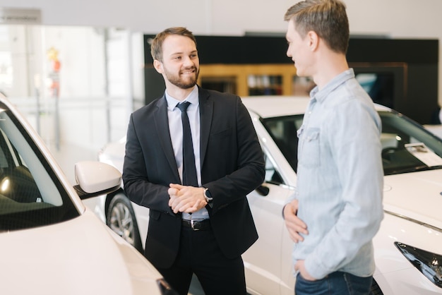 Il venditore di auto fiducioso che indossa un tailleur sta raccontando il nuovo modello di auto a un giovane che indossa abiti casual nella concessionaria di automobili. Concetto di scelta e acquisto di una nuova auto presso lo showroom.