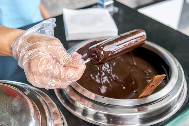 Il venditore in guanti di plastica immerge il gelato sul bastoncino nella cioccolata calda Processo di produzione di ghiaccioli