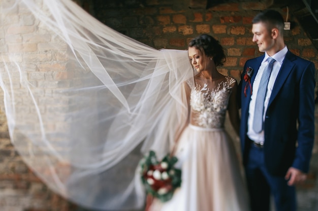 Il vento soffia via il velo della sposa mentre lei posa con lo sposo prima del muro di mattoni