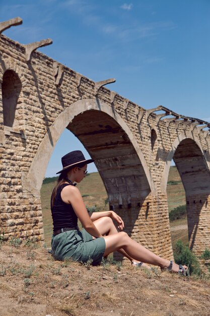 Il viaggiatore con un cappello nero si siede vicino al vecchio ponte di pietra distrutto in estate