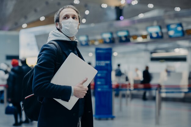 Il viaggiatore dell'uomo indossa una maschera medica monouso protettiva in aeroporto, ritorna dall'estero dove si diffonde il coronavirus, porta zaino, si prende cura della salute, protegge dai virus, ha posticipato il volo