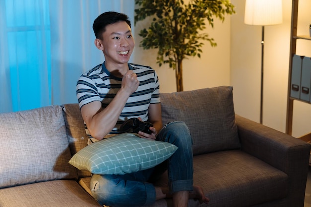 Il vincitore dell'uomo giapponese asiatico eccitato e felicissimo tiene il joystick sentendosi euforico con la vittoria del videogioco nel soggiorno a tarda notte. ragazzo felice guardare la tv celebrare la vittoria. maschio sorridente che si diverte la sera