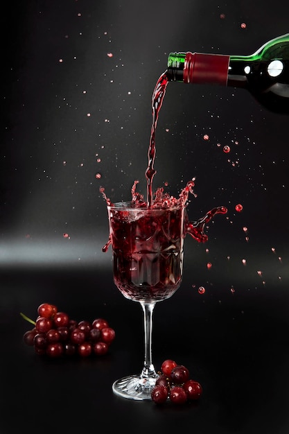 Il vino rosso viene versato in un bicchiere di vino su uno sfondo nero con uva e spruzzi