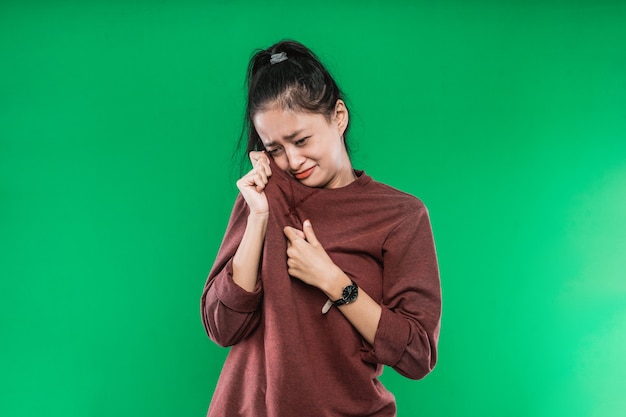 Il volto di un'espressione che piange di una giovane donna asiatica mentre si asciuga le lacrime con i vestiti isolati su sfondo verde