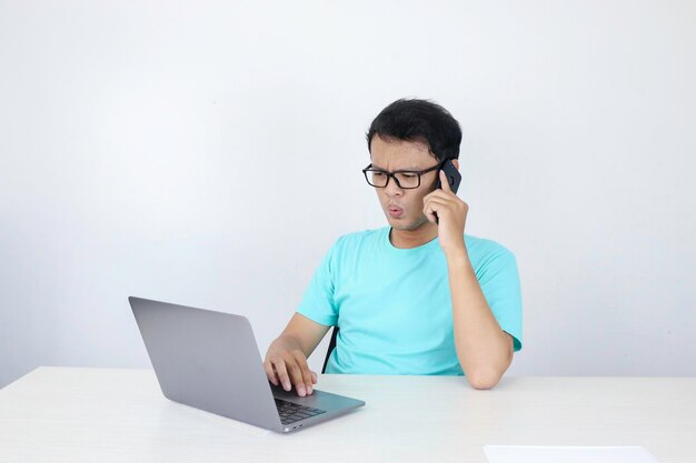 Il volto wow del giovane asiatico ha scioccato ciò che vede nel laptop quando chiama al telefono Uomo indonesiano che indossa una camicia blu