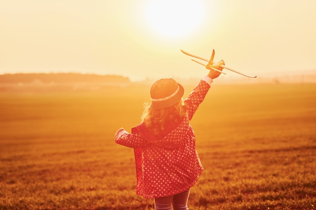 Illuminato dalla luce del sole arancione. La bambina sveglia si diverte con l'aereo del giocattolo sul bellissimo campo di giorno