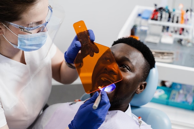 Illuminazione UV del riempimento del dente in fotopolimero per l'uomo africano in odontoiatria Dentista in occhiali protettivi tratta e rimuove la carie del paziente africano