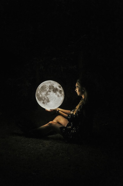 Illusione ottica di una donna che tiene la luna mentre è seduta sul campo di notte