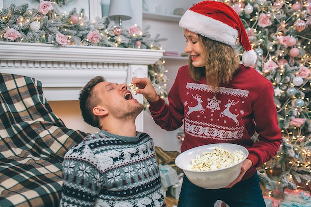 Immagine adorabile della giovane donna che sta e che alimenta il suo ragazzo. Tiene un pezzo di popcorn sopra la sua bocca. Guy tiene la bocca aperta. Il giovane si siede sul pavimento.