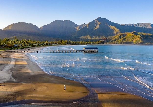 Immagine aerea all'alba al largo della baia di Hanalei e del molo sull'isola hawaiana di Kauai con un uomo in piedi da solo sulla spiaggia