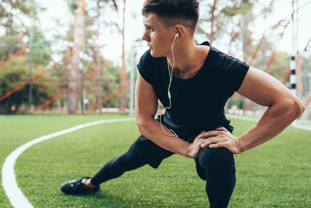 Immagine all'aperto di un giovane muscoloso che si allunga sullo sfondo della natura Uomo atleta che distoglie lo sguardo allungando la gamba e ascoltando la musica sugli auricolari Stile di vita sportivo e concetto di persone