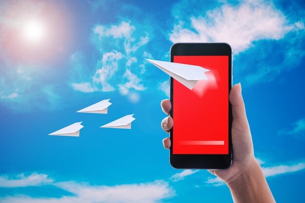 Immagine composita digitale di una persona che tiene in mano uno smartphone con un aereo di carta contro il cielo blu