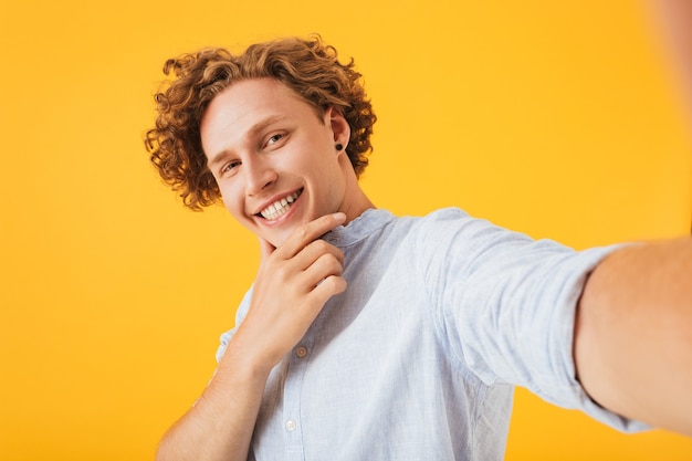 Immagine del bel ragazzo riccio prendendo selfie foto e toccando il mento, isolato su sfondo giallo