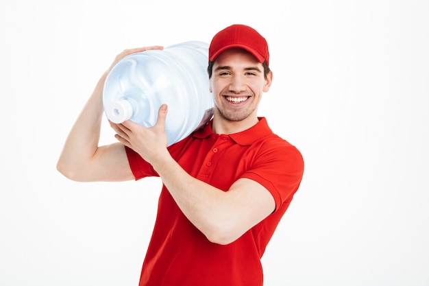 Immagine del corriere sorridente di consegna dell'acqua in bottiglia in maglietta rossa e carro armato di trasporto del cappuccio della bevanda fresca, isolato sopra spazio bianco