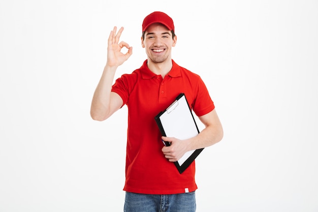 Immagine del fattorino ottimista in maglietta rossa e cappuccio che sorride e che mostra segno giusto mentre tenendo lavagna per appunti, isolato sopra spazio bianco