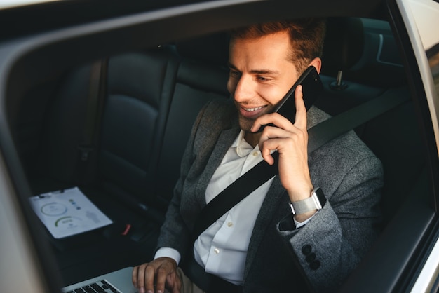 Immagine del giovane uomo regista in tuta parlando su smartphone e lavorando sul portatile, mentre torna seduto in auto di business class con cintura di sicurezza