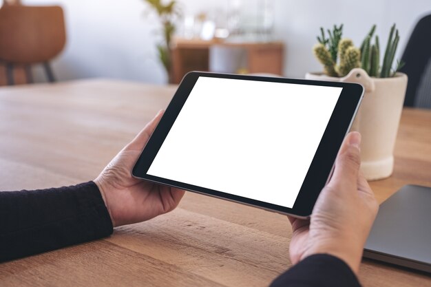 Immagine del mockup delle mani che tengono e utilizzando il tablet pc nero con schermo desktop bianco vuoto con il taccuino sulla tavola di legno in ufficio