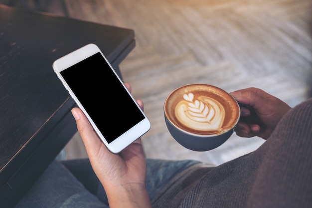 Immagine del modello delle mani che tengono un telefono cellulare bianco con uno schermo vuoto mentre beve il caffè