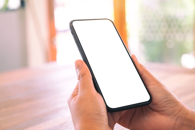 Immagine del modello delle mani della donna che tengono il telefono cellulare nero con lo schermo bianco in bianco sulla tavola di legno