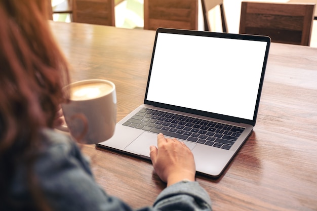 Immagine del modello di una donna che utilizza e tocca il touchpad del laptop con lo schermo del desktop bianco vuoto sulla tavola di legno mentre beve il caffè