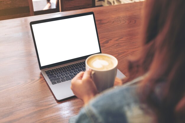 Immagine del modello di una donna che utilizza e tocca il touchpad del laptop con lo schermo del desktop bianco vuoto sulla tavola di legno mentre beve il caffè